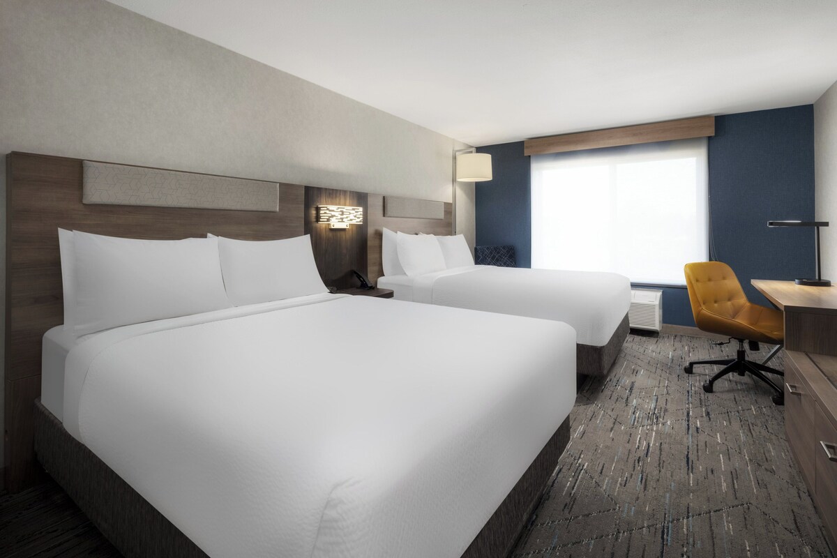 A hotel room with two beds, a desk, a chair, and a large window.