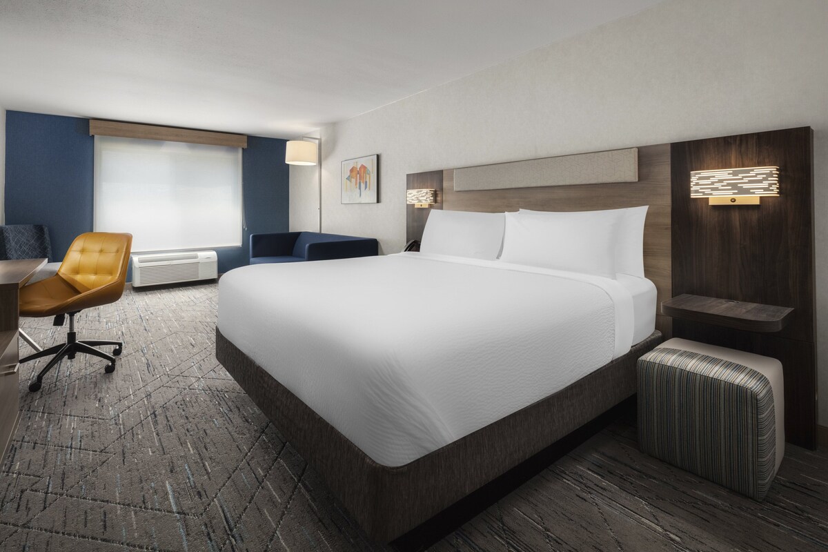 A hotel room with a large bed, a desk with a chair, a small ottoman, and a wall-mounted lamp.