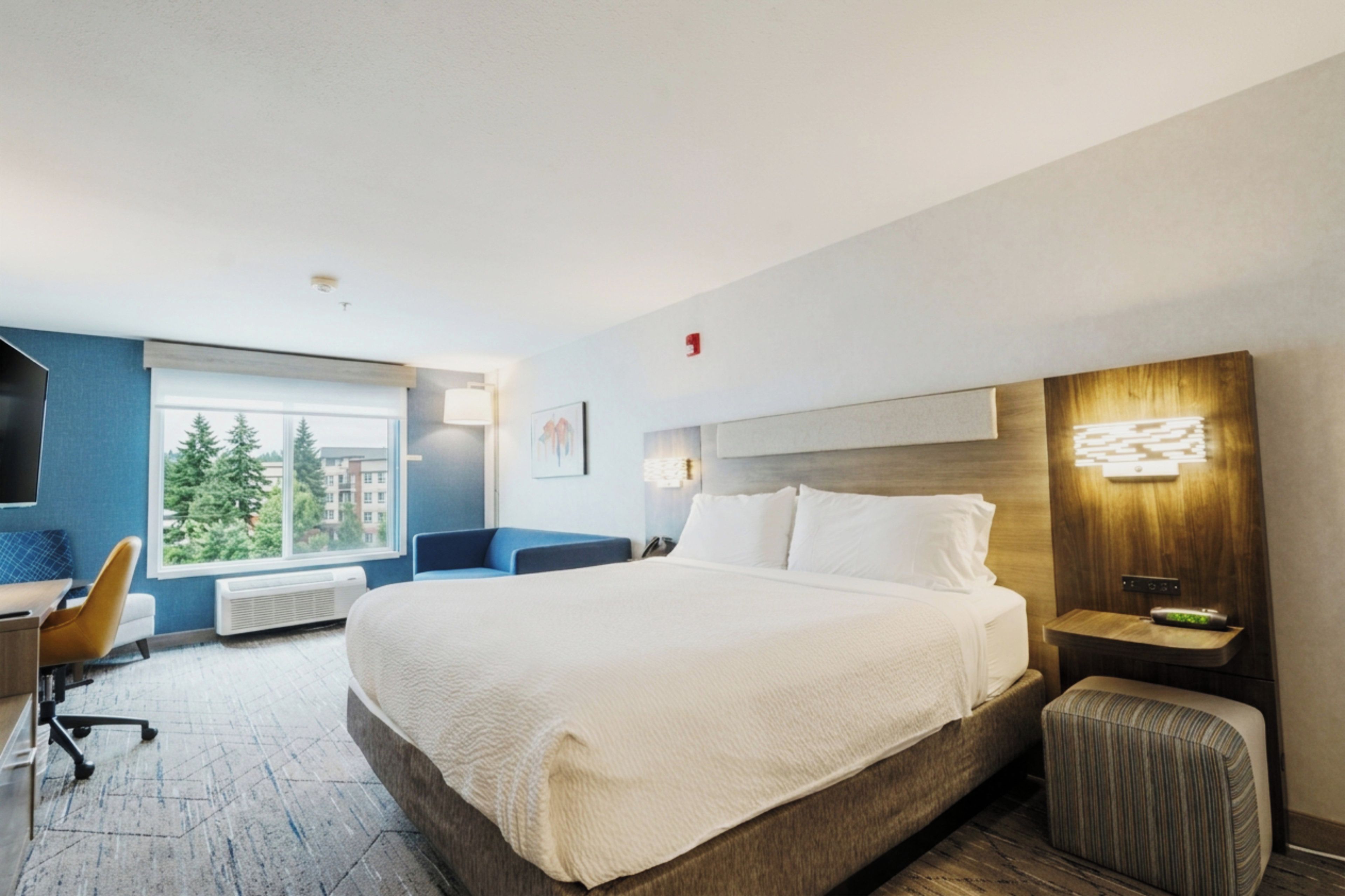 A hotel room with a large bed, a desk with a chair, a window with a view of trees, and a wall-mounted light fixture.