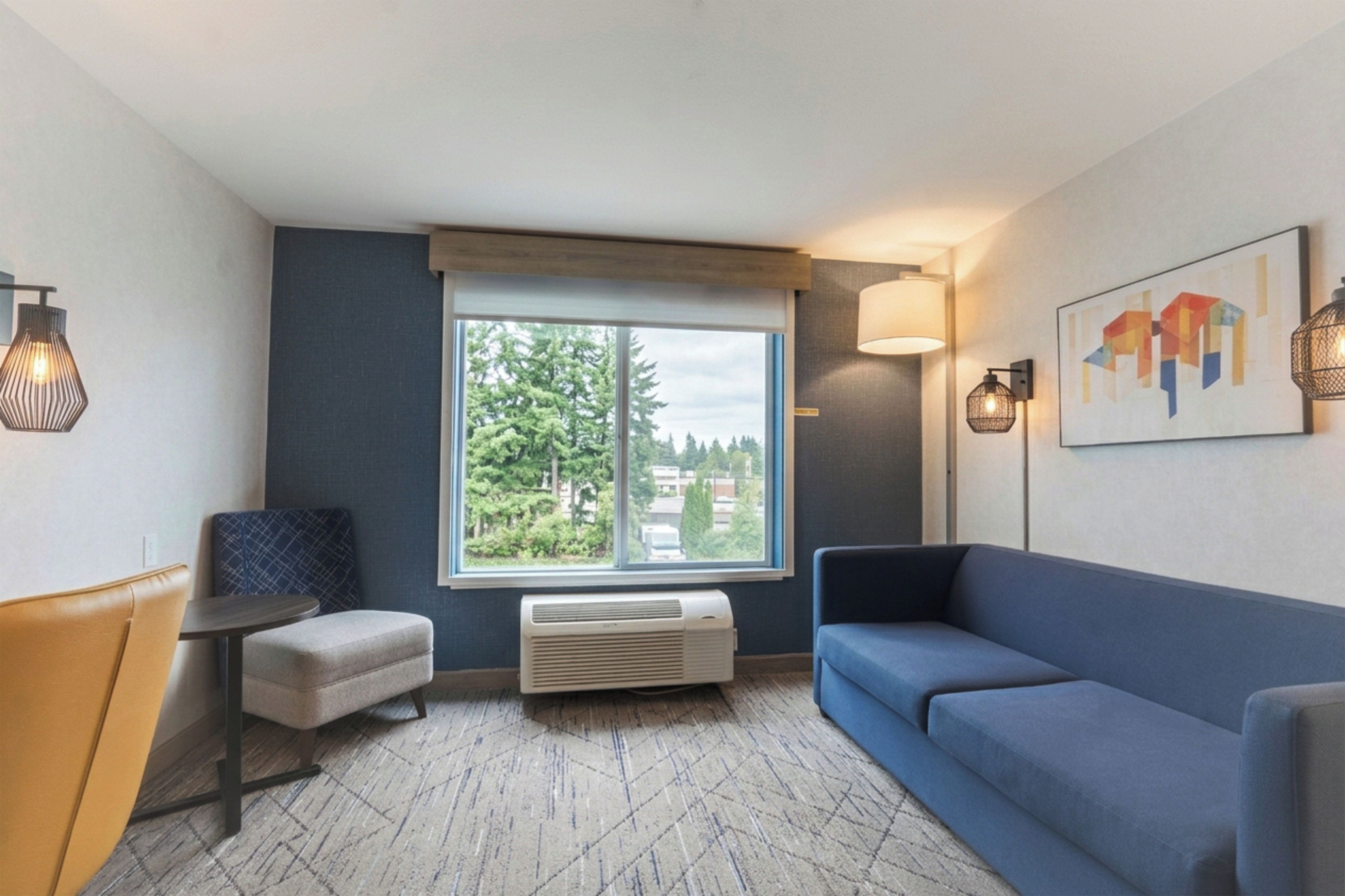 A hotel room with a large window, a blue sofa, a small table, and a chair.
