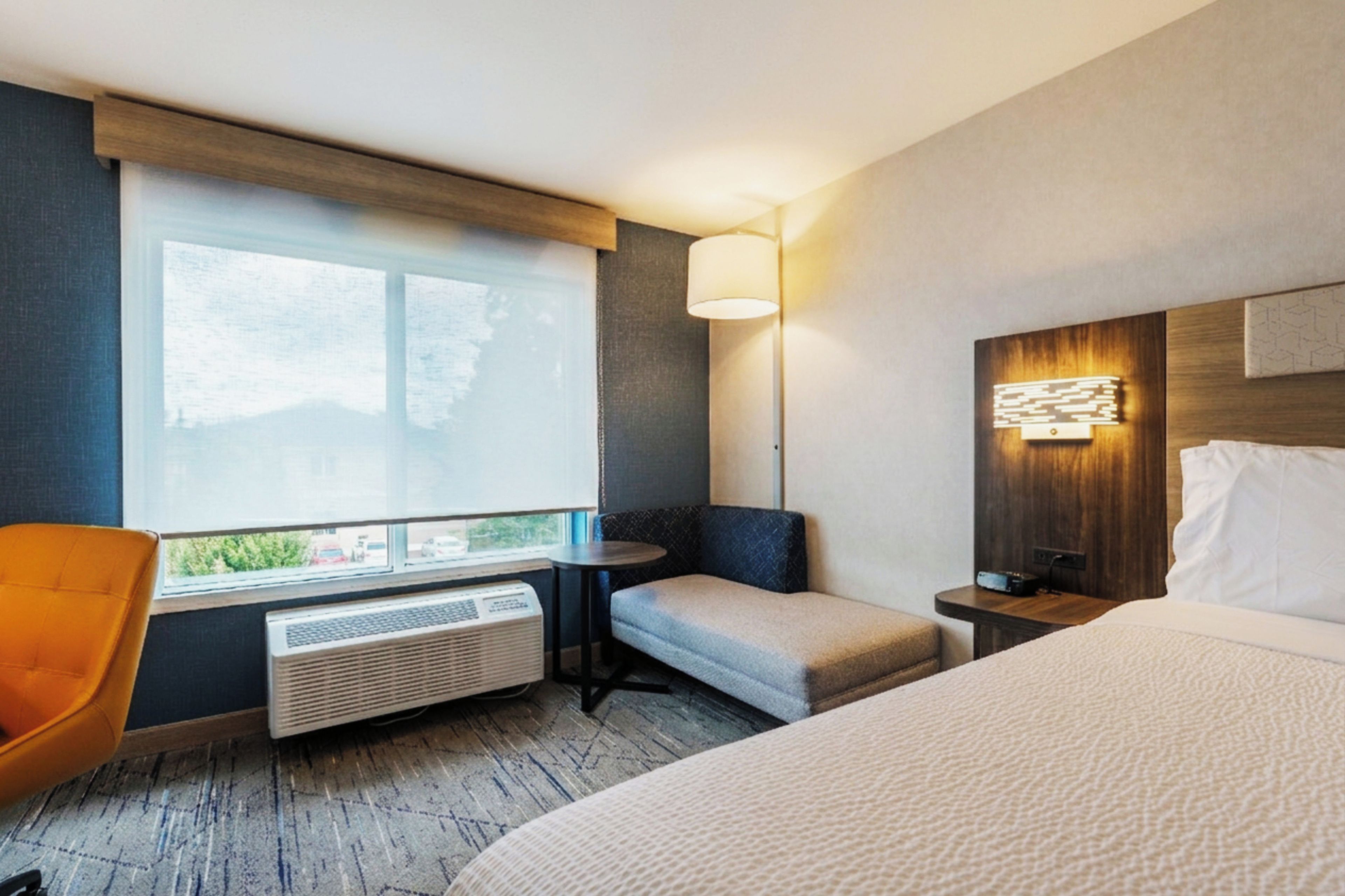 A hotel room with a bed, a seating area with a chair, a table, a lamp, and a heater.
