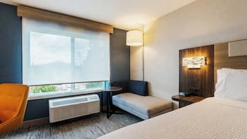 A hotel room with a bed, a seating area with a chair, a table, a lamp, and a heater.
