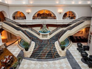 Un amplio vestíbulo de hotel con una escalera central, varias zonas de estar y una fuente decorativa.