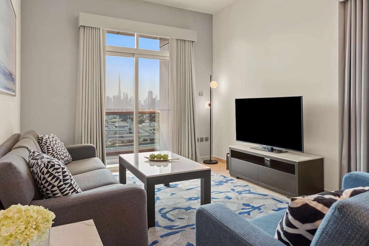 Una sala de estar moderna con sofá, mesa de centro, televisor y vistas a un horizonte urbano a través de amplios ventanales.