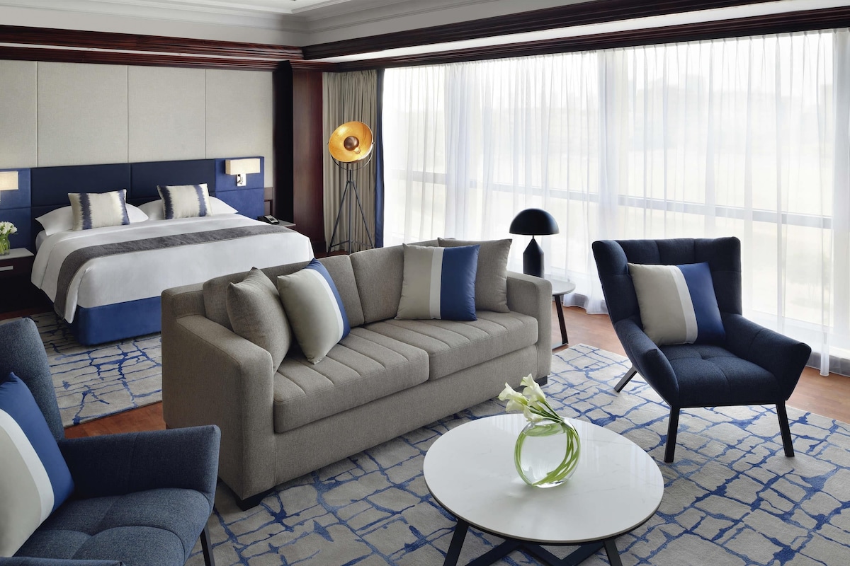 A hotel room with a bed, sofa, armchair, and a round table with a vase.