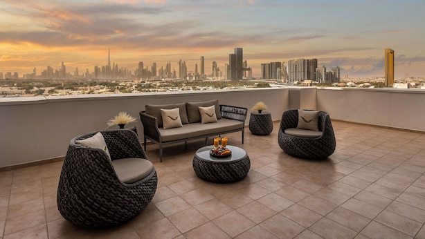 Una terraza en la azotea con un sofá, sillas y una mesa redonda, con vistas a un horizonte urbano al atardecer.