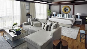 Una habitación de hotel moderna con sofá, reposapiés, una mesita con un frutero, cama, mesita de noche y un ventanal con cortinas.