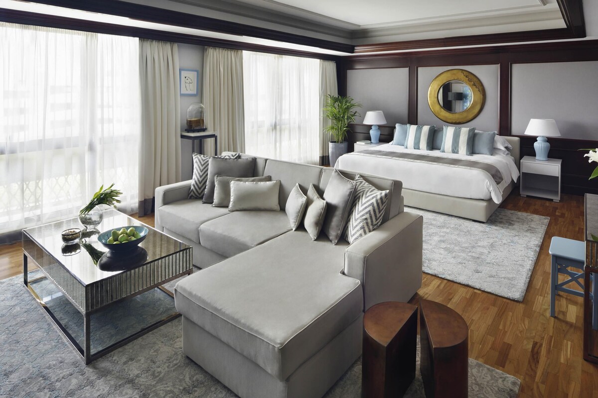 A modern hotel room with a sofa, ottoman, a small table with a bowl of fruit, a bed, a nightstand, and a large window with curtains.