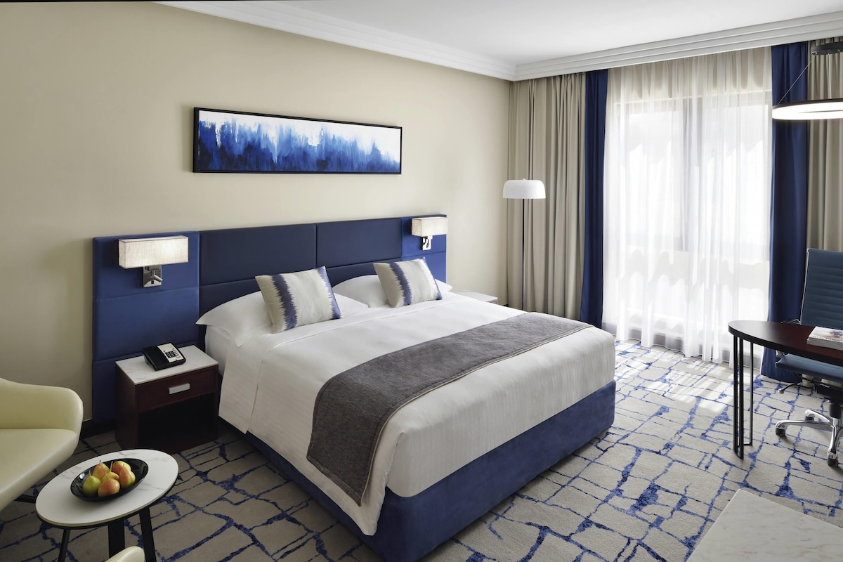 Habitación de hotel con una cama grande, un escritorio, una silla, una mesita con fruta y un cuadro en la pared.