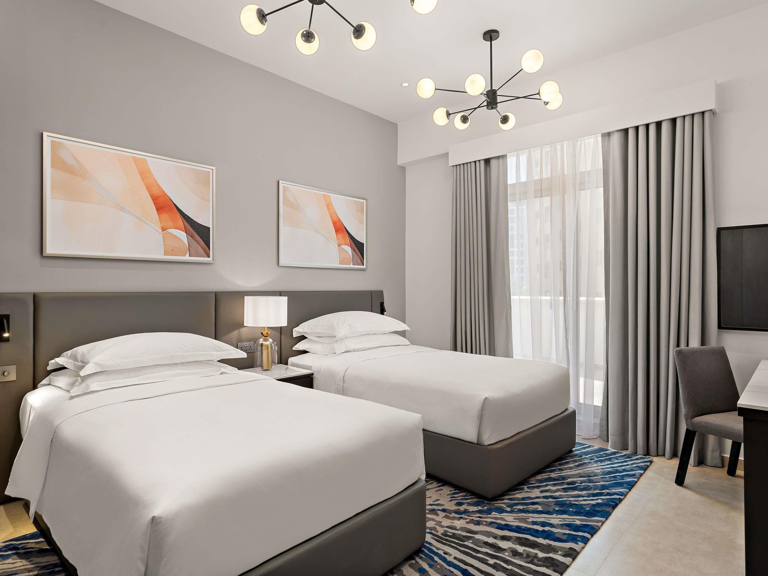 A hotel room with two beds, a desk, a chair, a large window with curtains, and a ceiling light fixture.