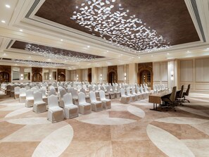 Un salón de conferencias amplio con filas de sillas blancas, una mesa larga con sillas y una lámpara en el techo.