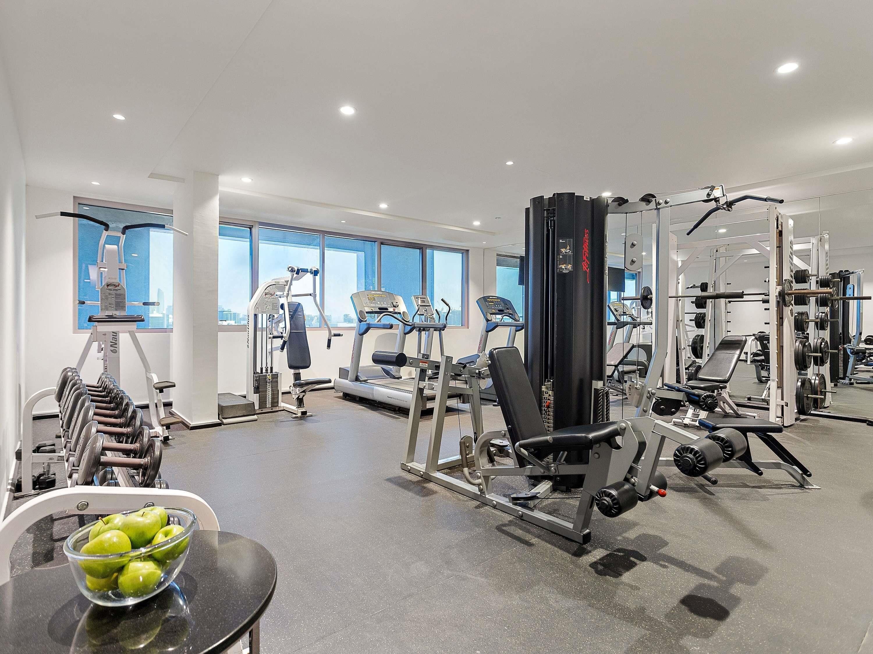A gym with various exercise equipment, a bowl of apples on a table, and large windows.