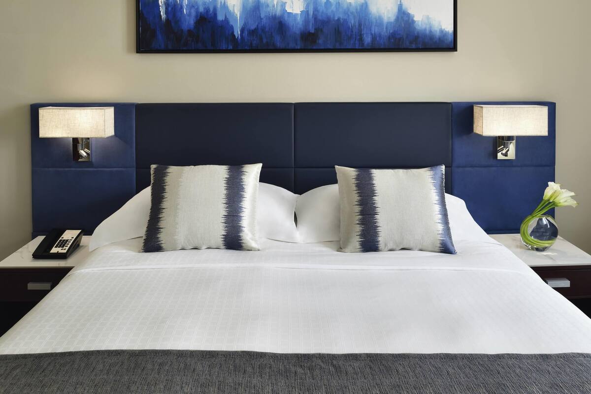 Un cuarto de hotel con una cama grande, dos lámparas de noche, un control remoto y un cuadro en la pared.