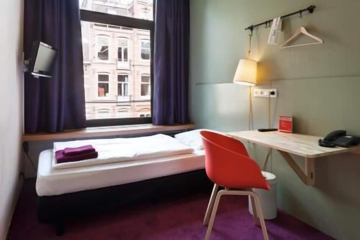 A hotel room with a bed, a desk, a chair, a lamp, a phone, and a window with curtains.