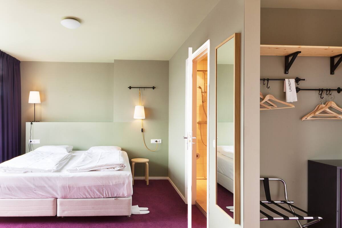A hotel room with a bed, a small stool, a mirror, a towel rack, and a wardrobe.