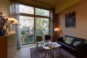A modern living room with a large window, a sofa, two chairs, a table with a candle, and a painting on the wall.