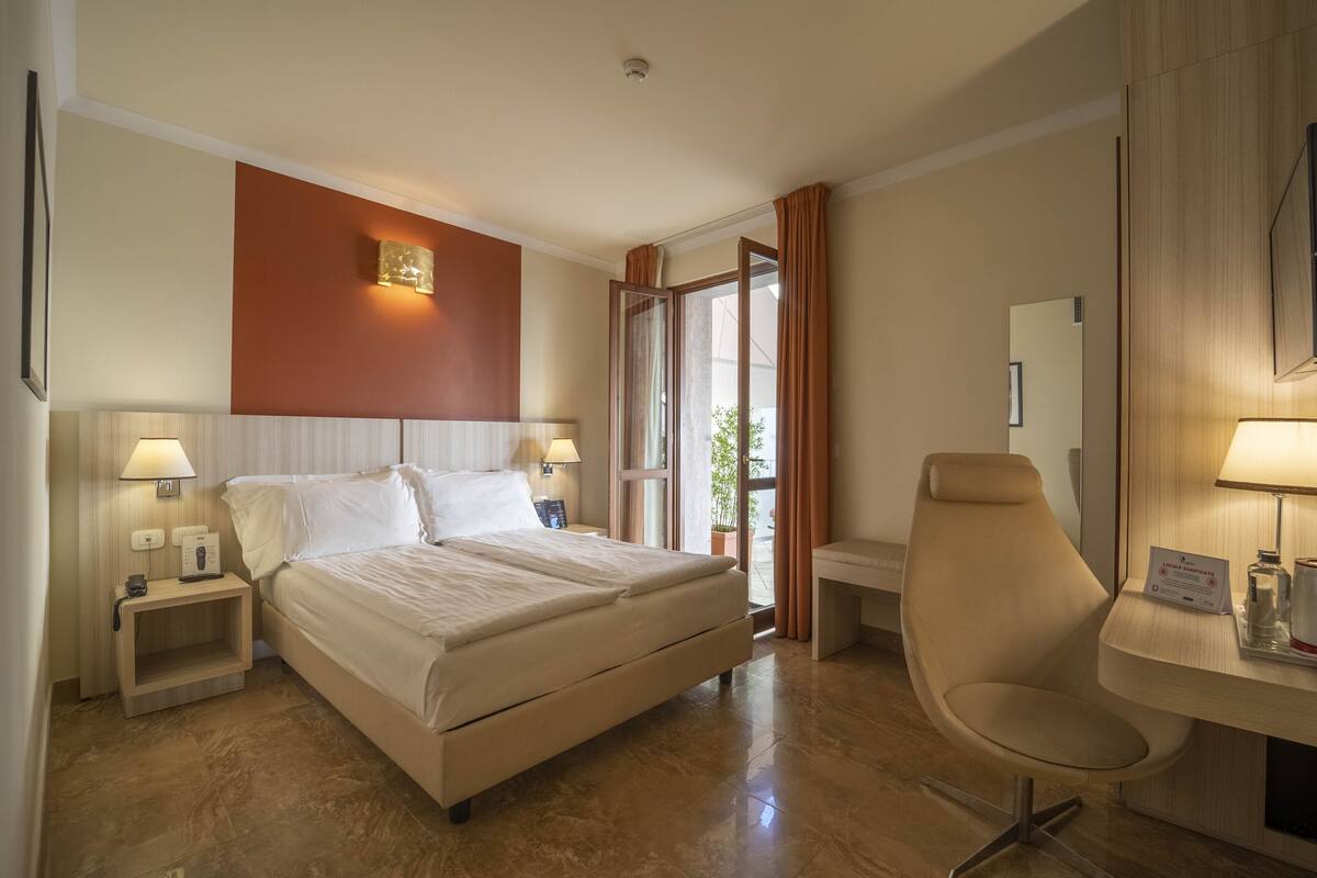 A hotel room with a bed, bedside tables, a desk, a chair, and a large window.