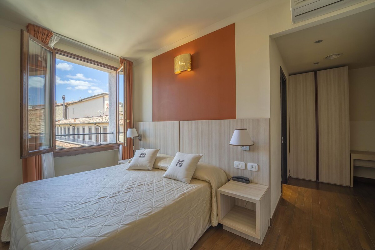 A hotel room with a bed, bedside tables, a lamp, a view of buildings outside, and a wardrobe.