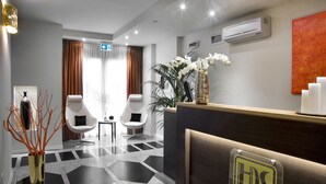 A hotel reception area with a modern design, featuring a reception desk, two white chairs, a vase with white flowers, and a geometric patterned floor.