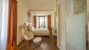 A modern hotel room with a bed, a chair, a desk, and a glass shower enclosure.
