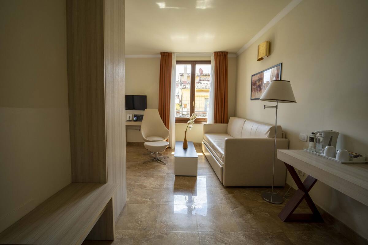 A modern living room with a sofa, armchair, coffee table, and a lamp. There is a TV and a window with curtains.