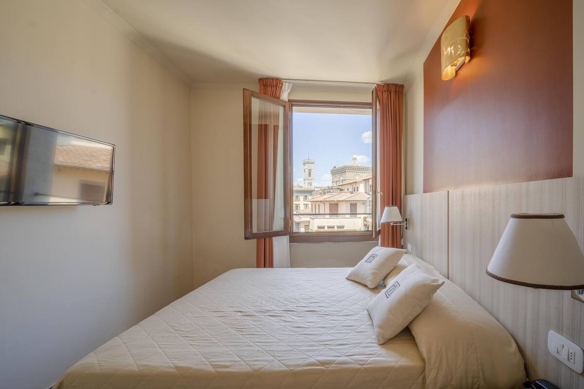 A hotel room with a bed, a TV mounted on the wall, a window with curtains, and a lamp on the bedside table.