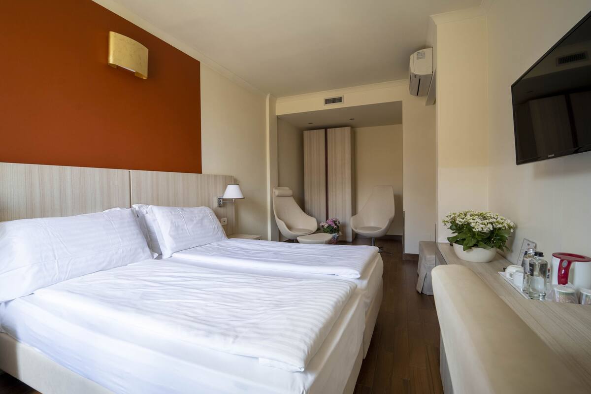 A hotel room with two beds, a TV, a desk with a vase of flowers, and a seating area with chairs.