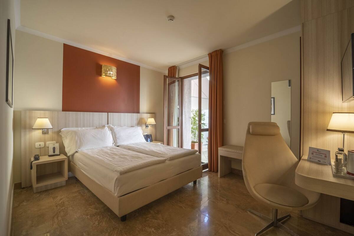 A hotel room with a bed, bedside tables, a desk, a chair, and a large window.