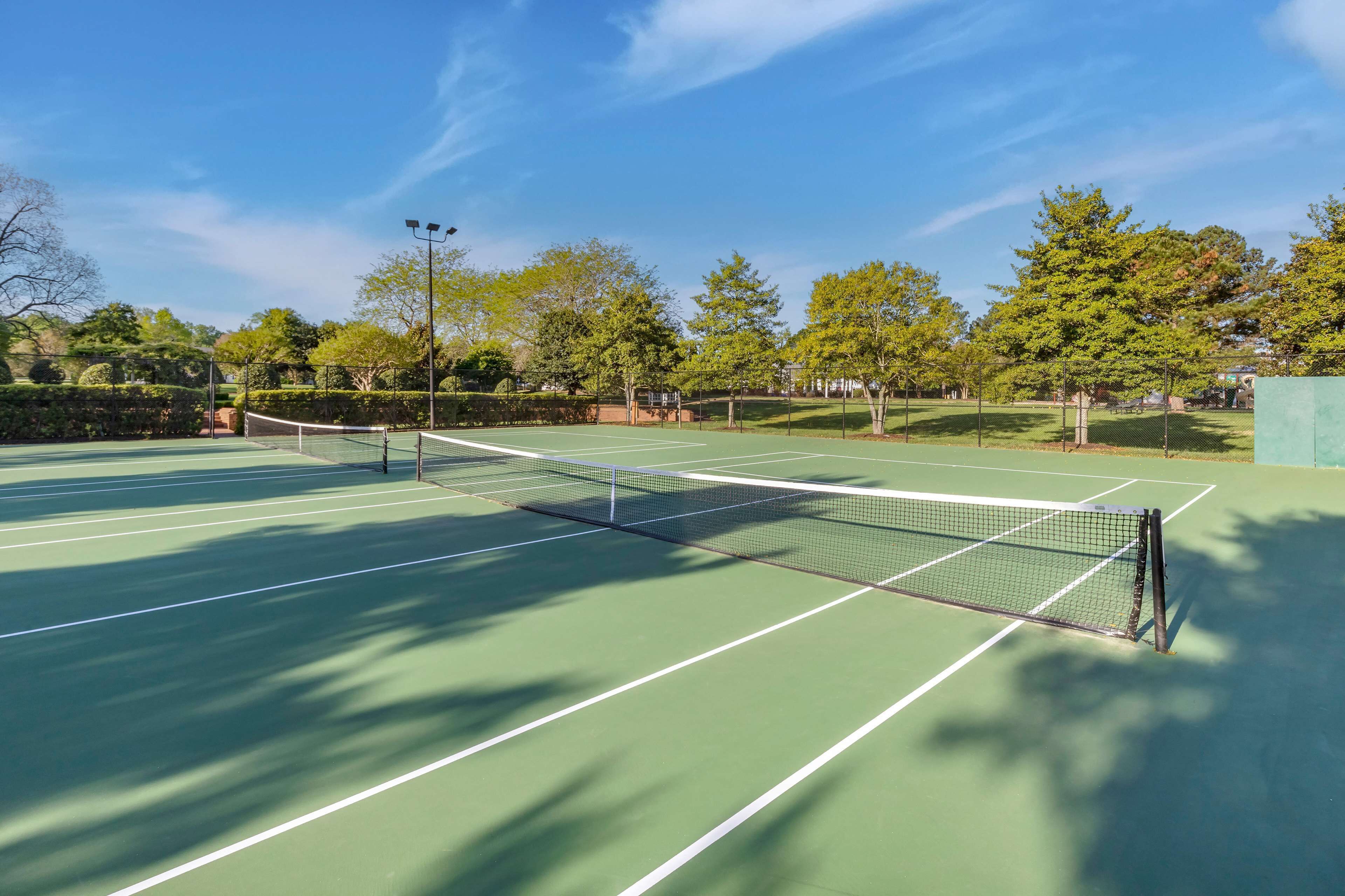 Una cancha de tenis al aire libre con red, rodeada de árboles y un cielo despejado.