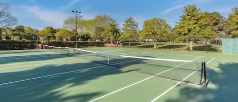 An outdoor tennis court with a net, surrounded by trees and a clear sky.