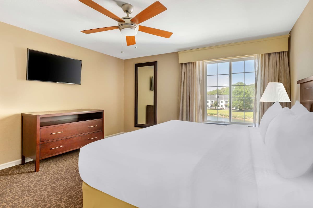 Ein Hotelzimmer mit einem Bett, Deckenventilator, Flachbildfernseher, Kommode, Spiegel, Fenster mit Vorhängen und einer Lampe.