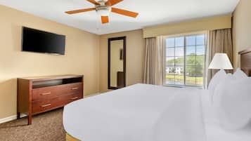 A hotel room with a bed, a ceiling fan, a flat-screen TV, a dresser, a mirror, a window with curtains, and a lamp.