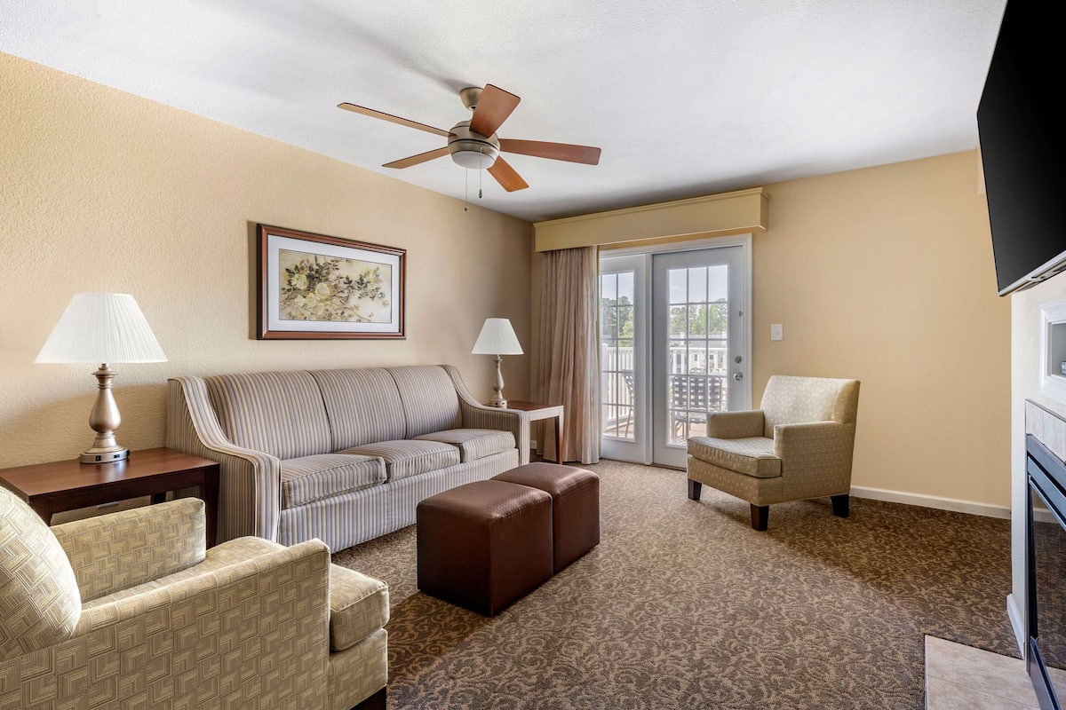 Ein Hotelzimmer mit einem gestreiften Sofa, einem braunen Ottomanen, einem beige farbenen Sessel, einem Deckenventilator und einem großen Fenster mit Vorhängen.