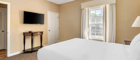 A hotel room with a bed, a flat-screen TV mounted on the wall, a small table with a lamp, and a window with curtains.