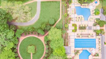 An aerial view of a hotel with a central swimming pool, landscaped gardens, and a winding road.