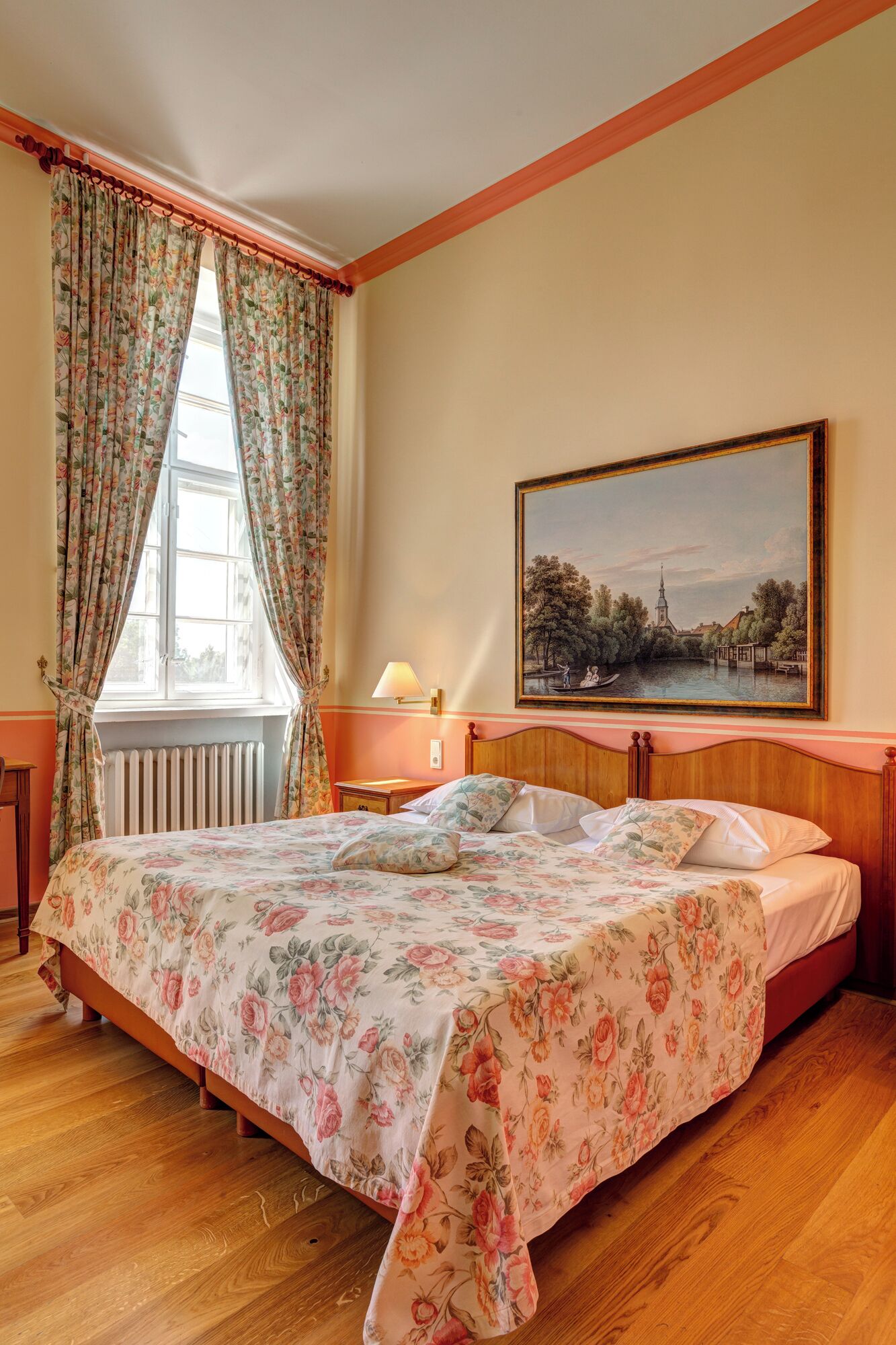 A bedroom with a wooden bed, floral bedspread, a window with curtains, a nightstand, a lamp, and a painting on the wall.