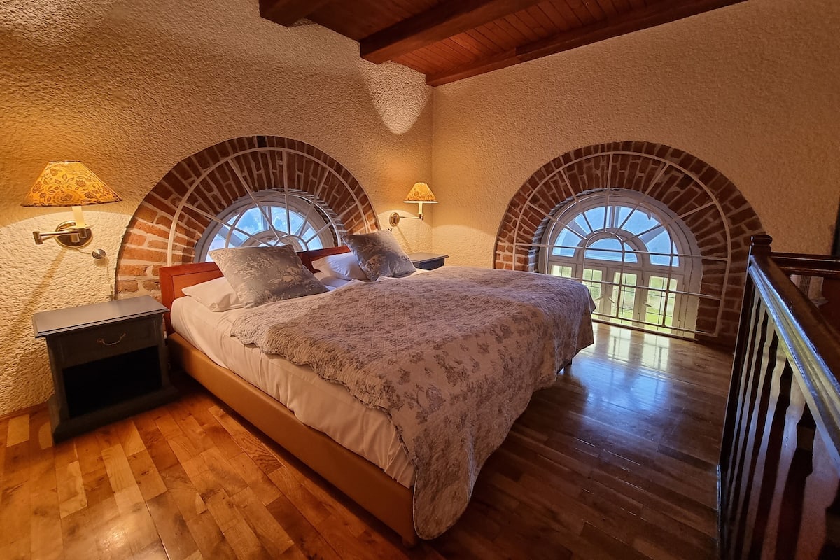 Un dormitorio con una cama de madera, dos lámparas fijadas a la pared y vistas a través de ventanales en arco.