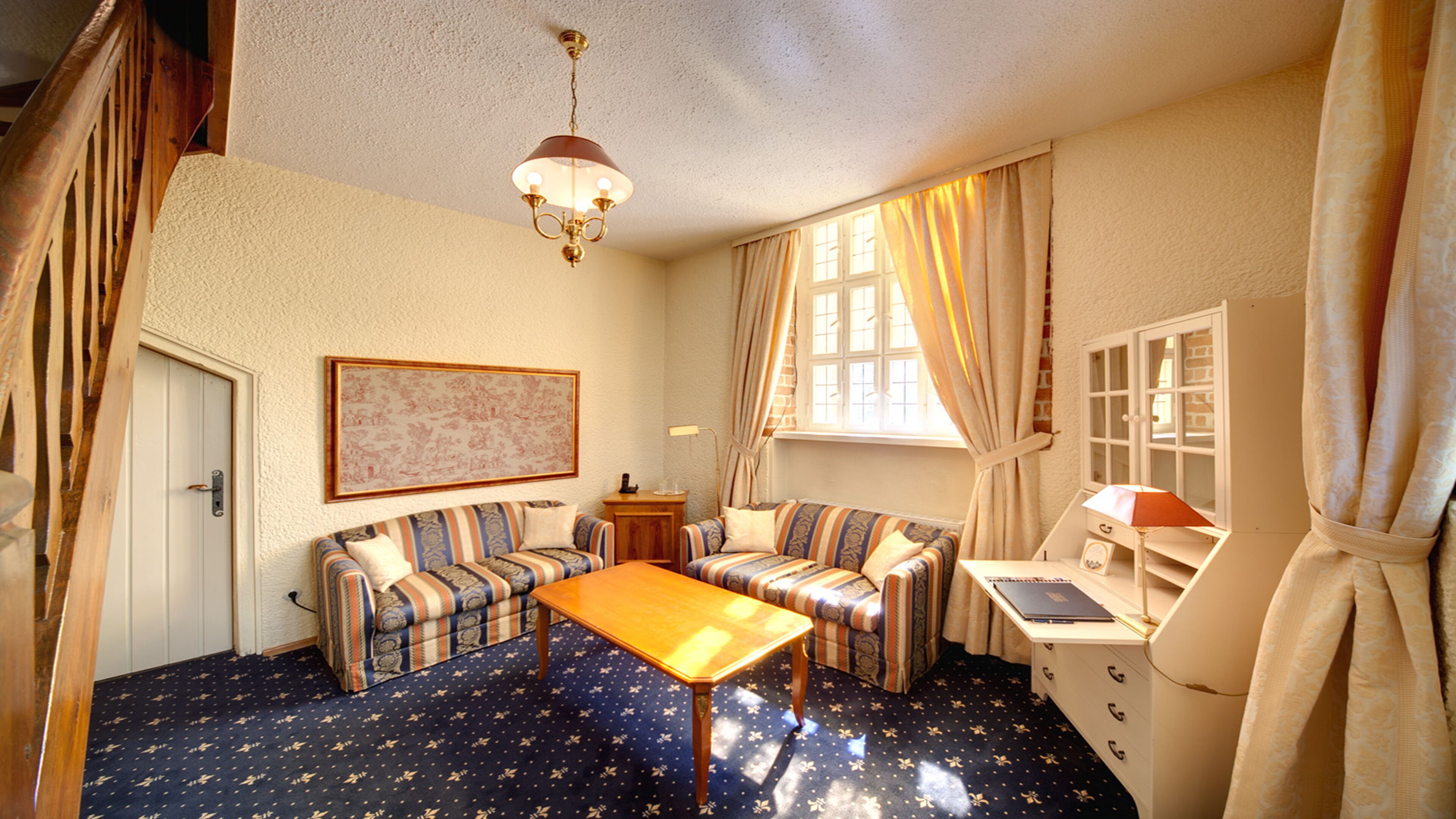Ein gemütliches Zimmer mit einer Sofagarnitur, einem Holztisch, einem Schreibtisch mit Lampe und einem fenster mit Vorhängen.