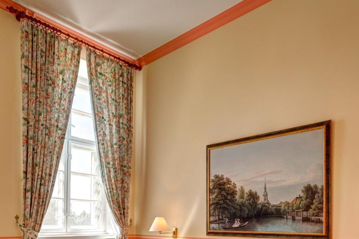 Un dormitorio con una cama de madera, ropa de cama floral, una ventana con cortinas, una mesita de noche, una lámpara y un cuadro en la pared.