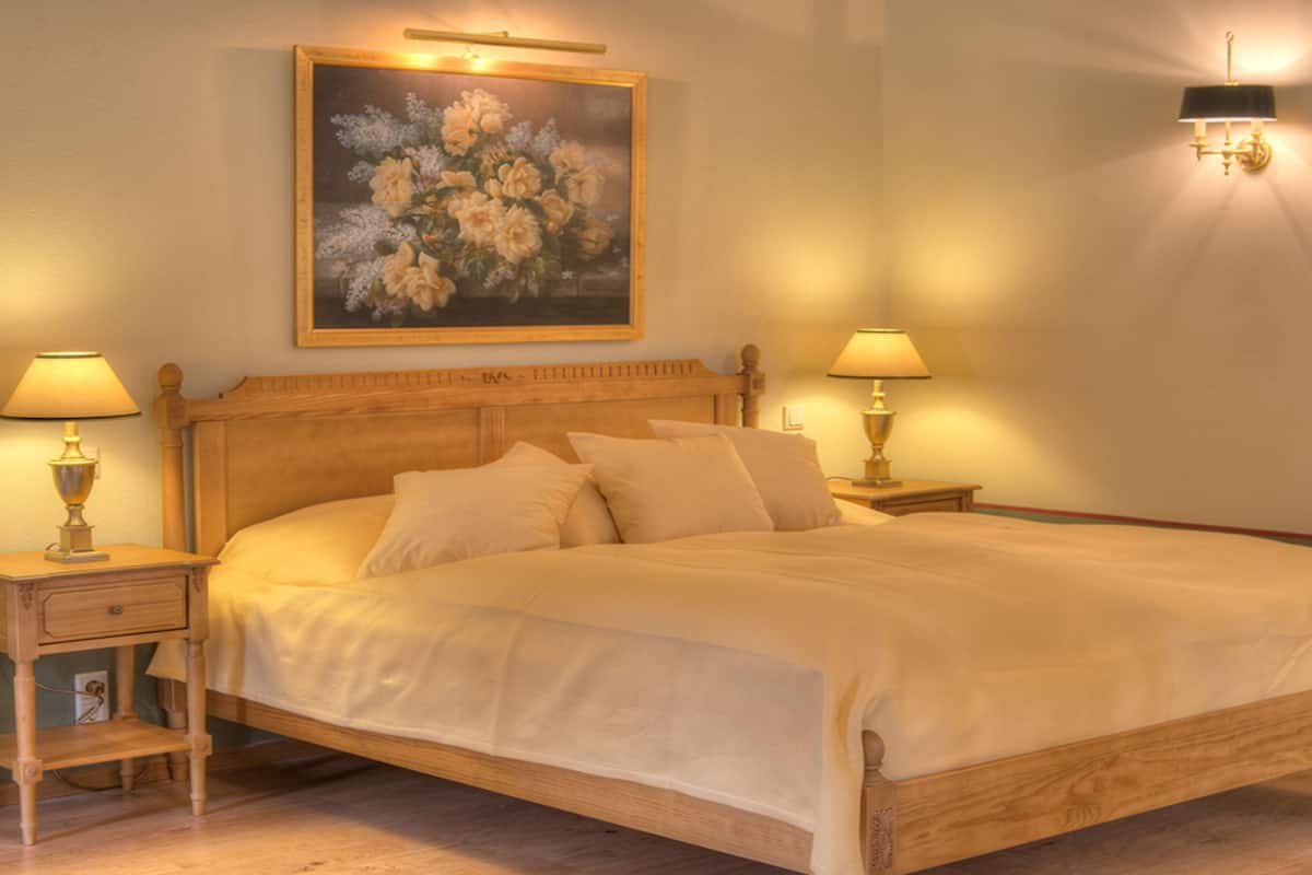Un dormitorio con una cama de madera, dos mesitas de noche con lámparas, una mesita de noche adicional con una lámpara y un cuadro en la pared.