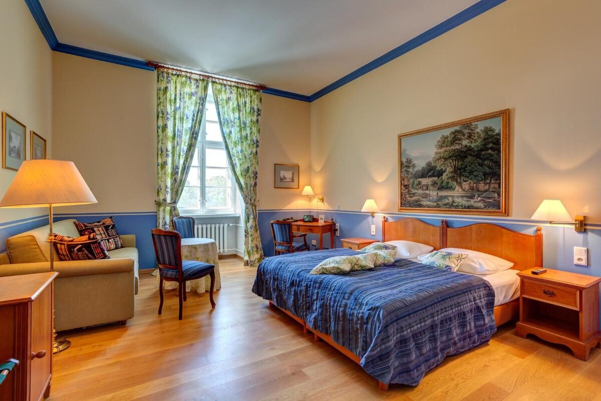 Ein Hotelzimmer mit Bett, Schreibtisch, Sessel, Sofa und einem Fenster mit Vorhängen.