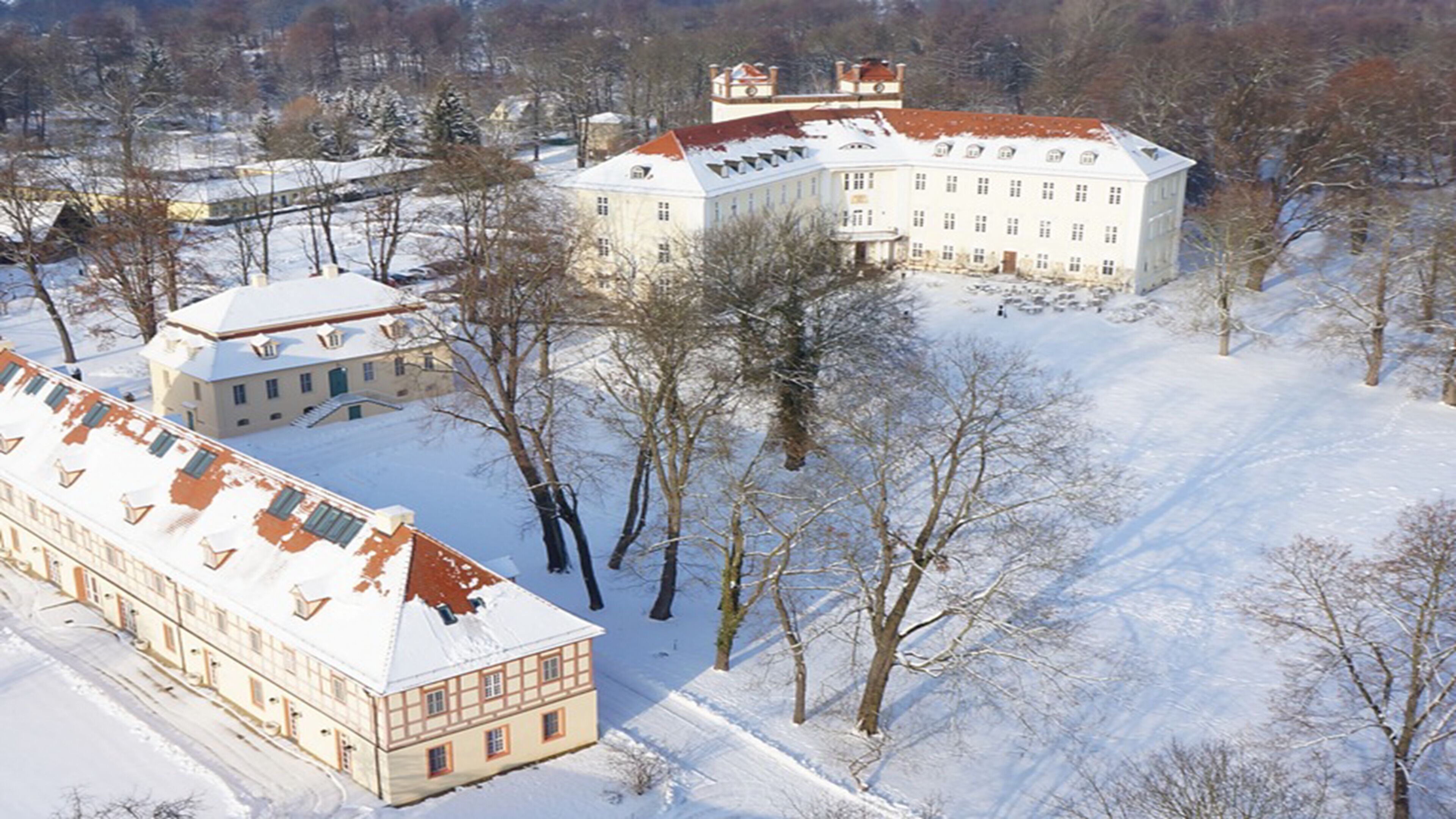 A large, historic building with multiple wings and a symmetrical facade, surrounded by snow-covered trees and a clear sky.