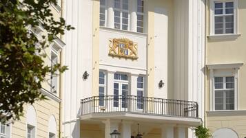 Ein historisches Gebäude mit einem zentralen Eingang, einem Balkon mit schmiedeeisernen Geländern und einem Wappen über dem Eingang.