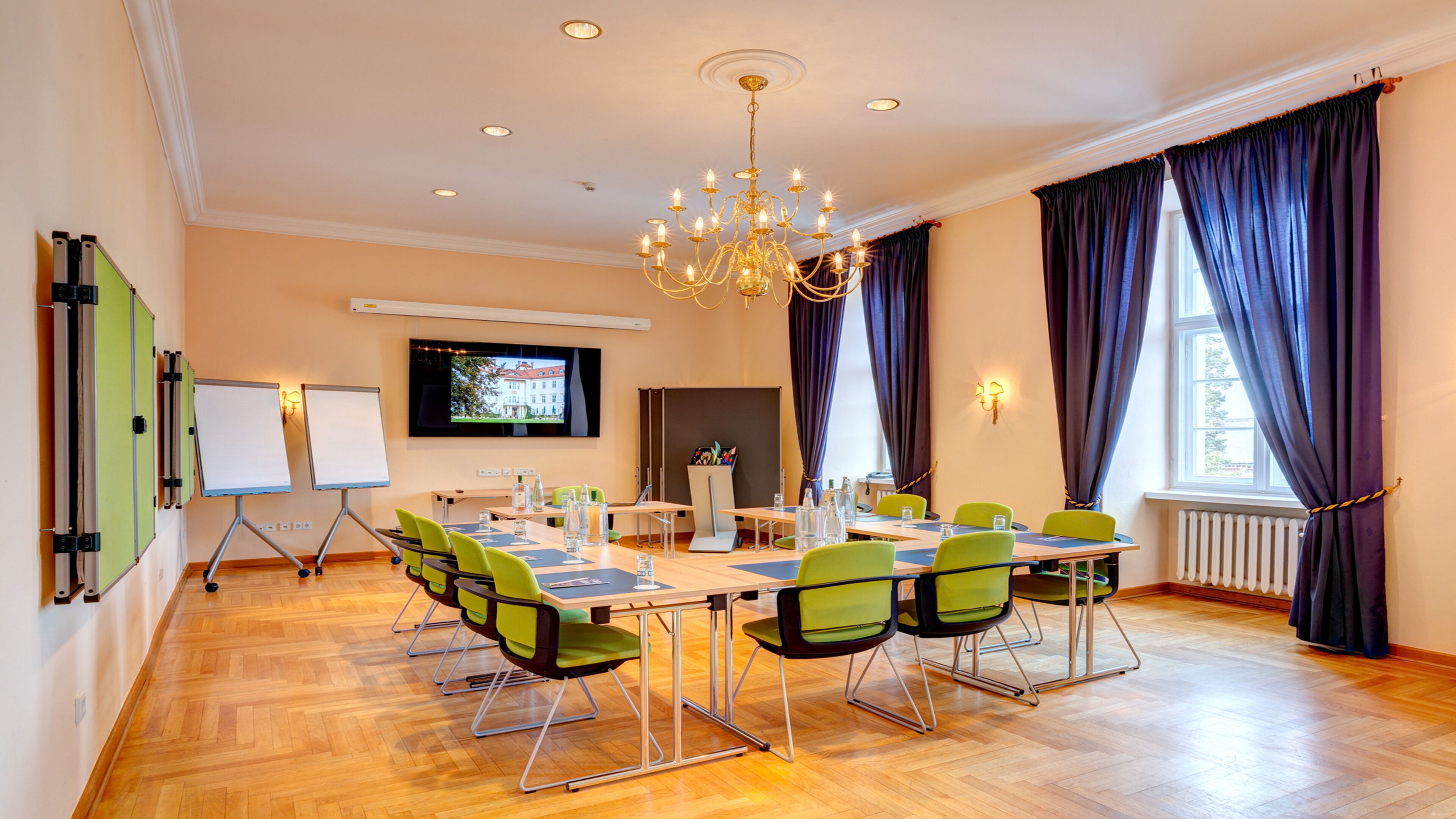 A conference room with a large table, green chairs, a chandelier, a whiteboard, and a TV on the wall.