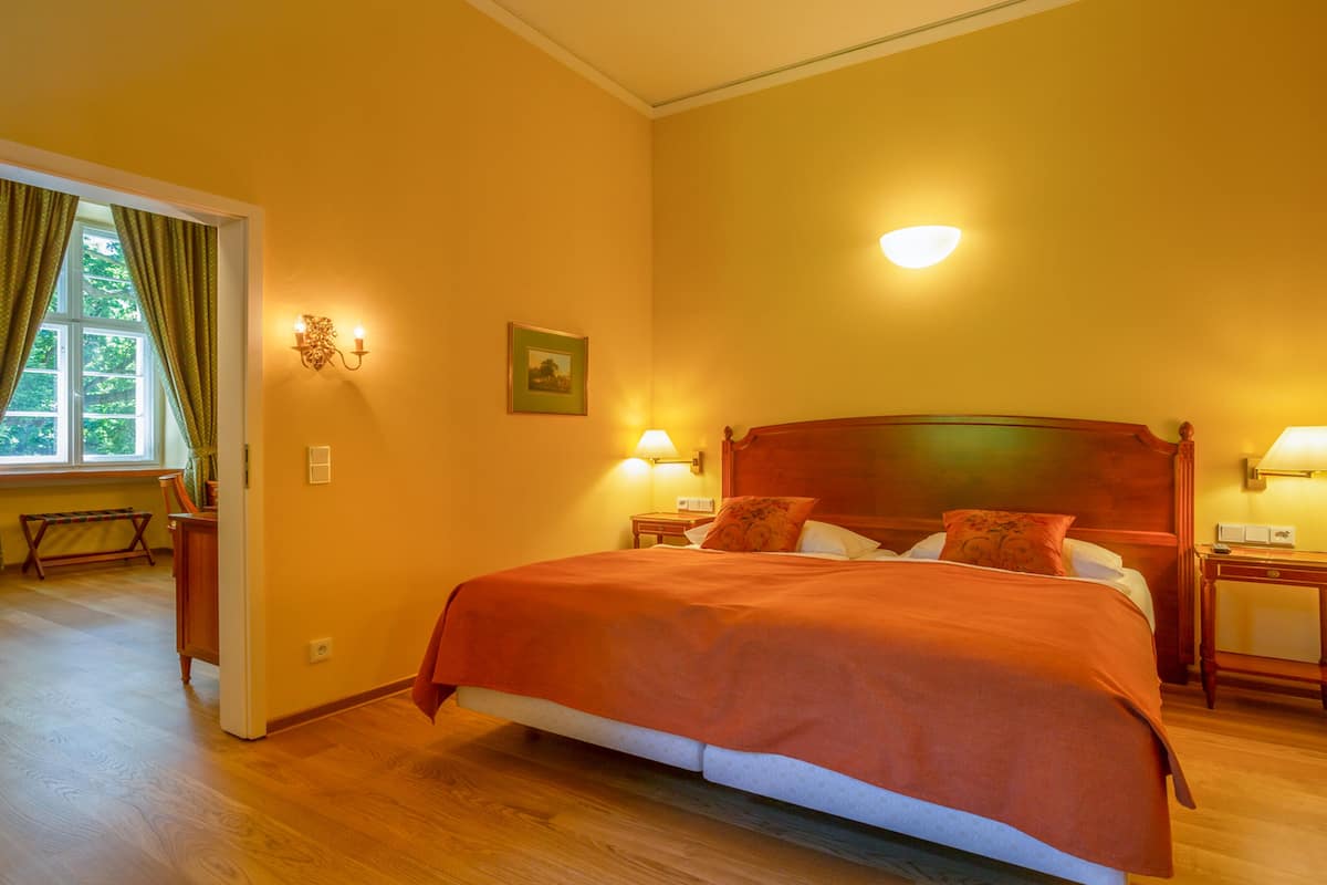 Habitación de hotel con una cama grande, dos mesitas de noche con lámparas, un cabecero de madera, una ventana con cortinas y una pequeña mesa con una silla.