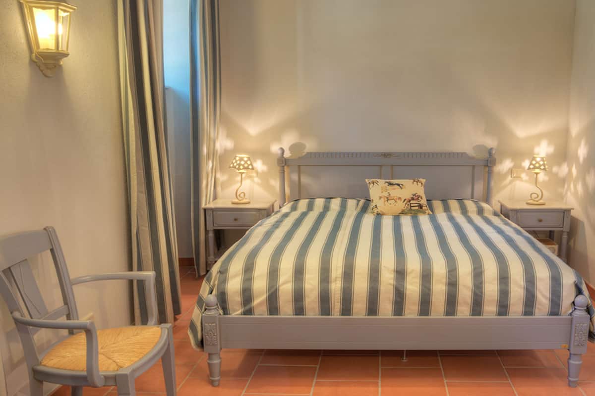 Un dormitorio con una cama, dos mesitas de noche, una silla y una lámpara de pared.