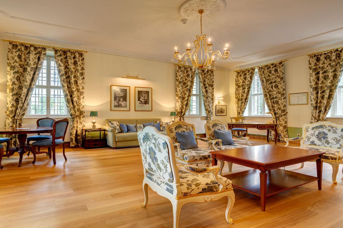 Una habitación amplia con piso de madera, un candelabro, cortinas con motivos florales y un área de comedor con sillas y una mesa.