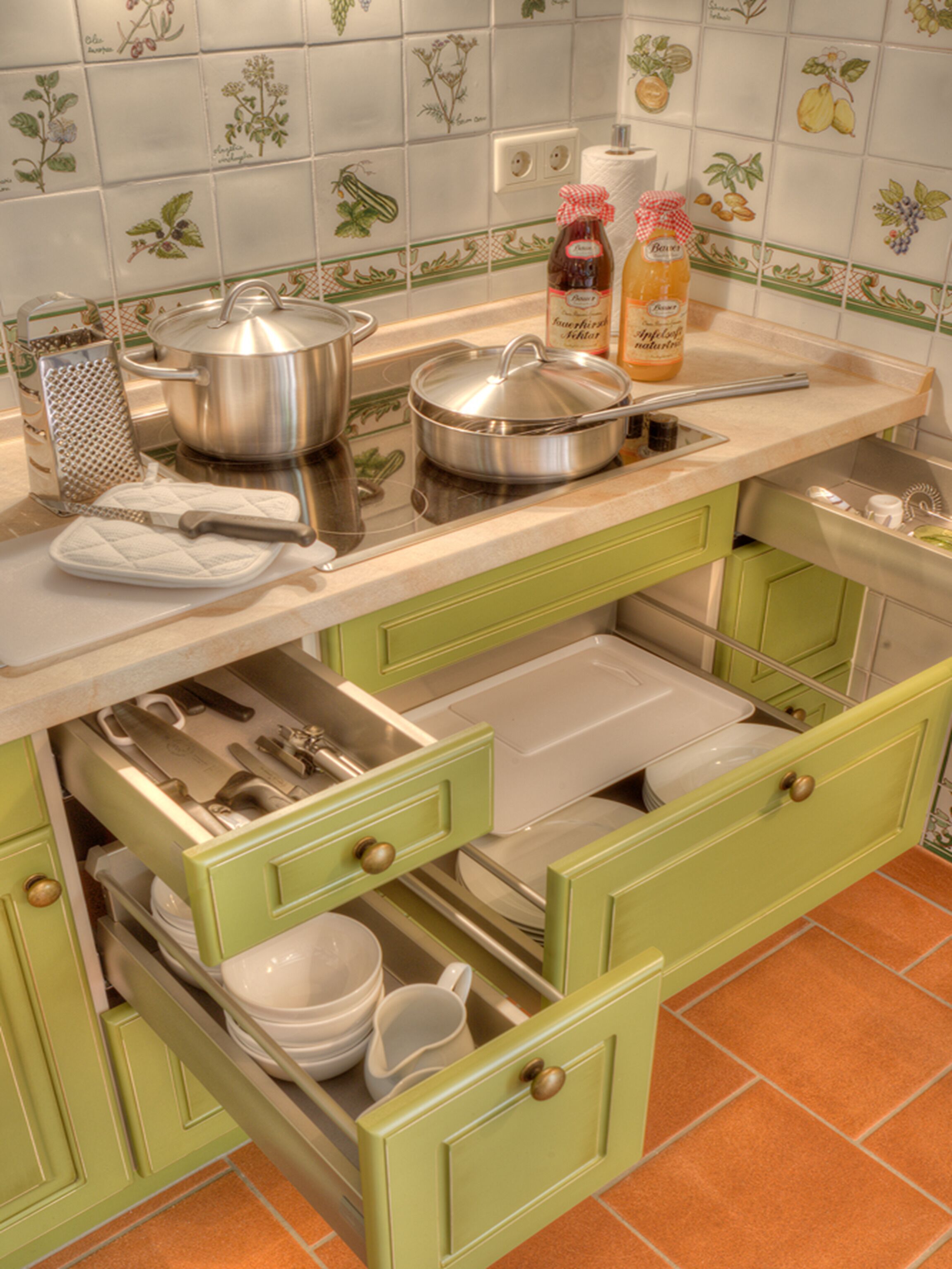 Una cocina con gabinetes verdes, una encimera con ollas y botellas, y un salpicadero de azulejos con motivos de frutas.