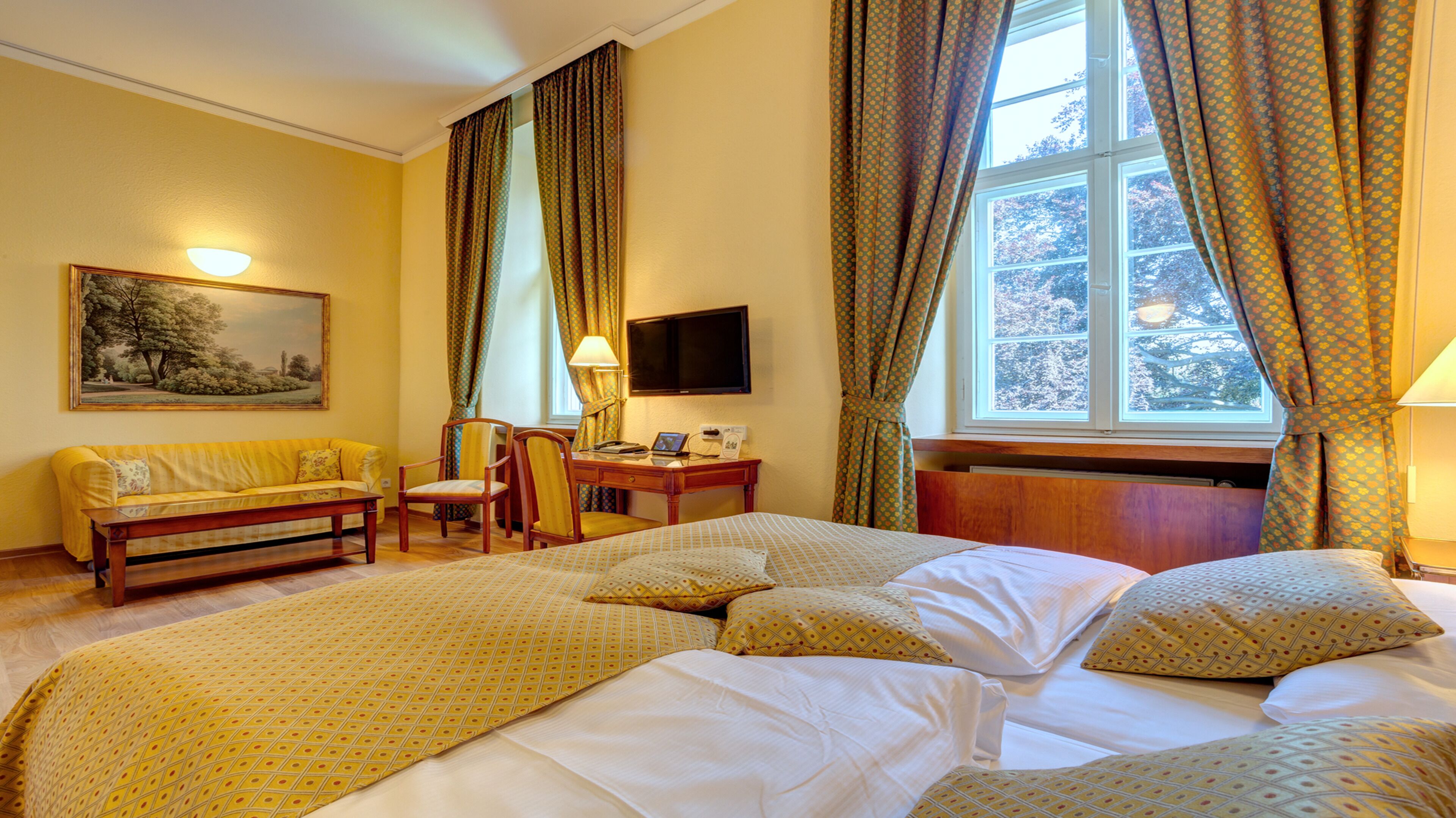 A hotel room with a large bed, a sofa, a desk with a chair, a television, and a painting on the wall.