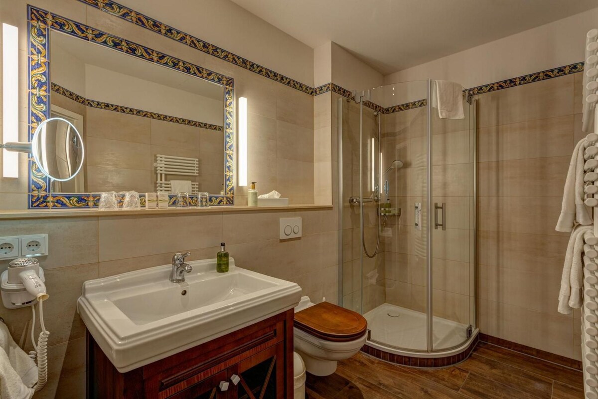 Une salle de bain avec un lavabo blanc, des toilettes en bois, une cabine de douche en verre et un miroir orné de carreaux décoratifs.